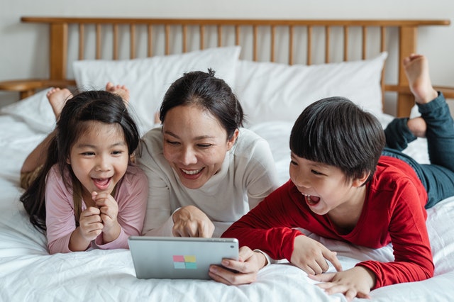 Mutter mit 2 Kindern im Bett schauen gemeinsam etwas Lustiges auf dem Tablet.